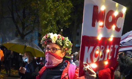 A woman wearing a crown of flowers and a scarf covering her face holds up a sign in the colours of the Polish flag with words in Polish